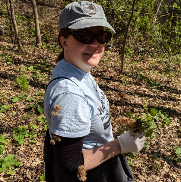A woman smiling at the camera while holding garlic mustard, with burrs stuck on her shirt.